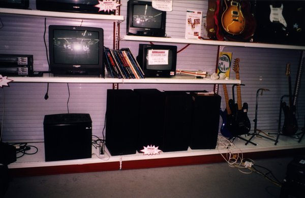 Rayonnage de magasin avec téléviseurs, enceintes et guitares électriques.