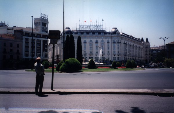 Homme debout face à un poteau devant un rond-point avec bosquets et fontaines.