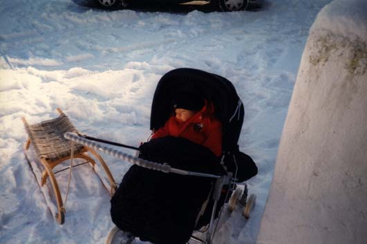 Enfant en anorak rouge endormi dans une poussette à l’arrêt sur un sol enneigé près d’une luge en bois.