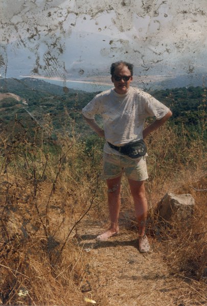 Homme debout en tee-shirt blanc et short, avec lunettes de soleil, sur un sentier avec végétations sèches, mer et bord côtier en arrière-plan. Image avec salissures.