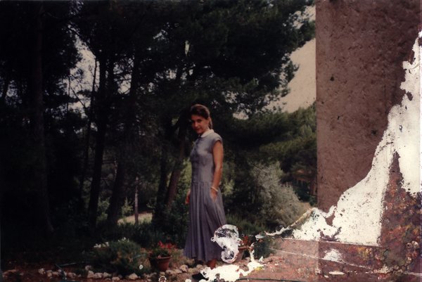 Jeune femme de trois-quart en robe longue gris-bleu debout sur le perron d’une maison, arbres en arrière-plan. Image abîmée.