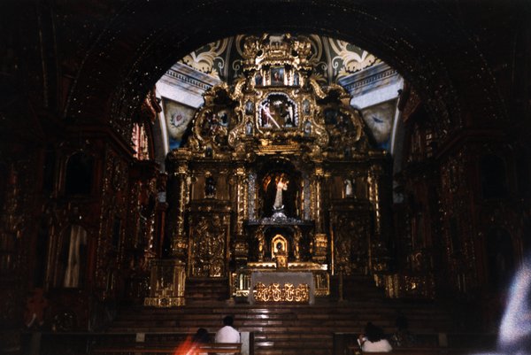 Chœur d’église et autel sculptés de style baroque, bancs et fidèles en avant-plan.
