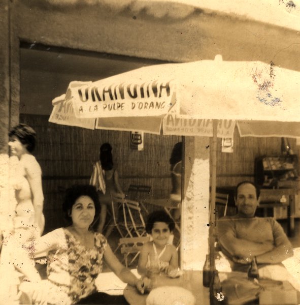 Homme torse nu avec femme et enfant installé sous un parasol Orangina à une terrasse de café. Noir et blanc.