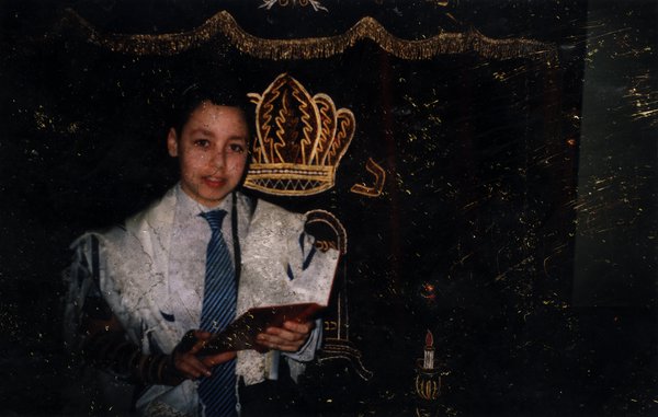 Enfant en châle de prière, un livre ouvert à la main, devant une tenture avec inscriptions en hébreu.