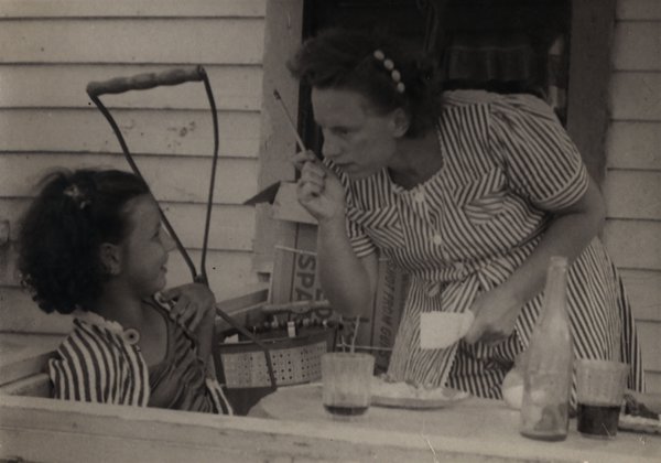 Femme en robe penchée sur une petite fille assise devant une table avec des couverts et une bouteille.