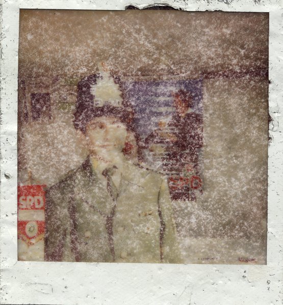 Jeune homme souriant en costume et chapeau devant affiche et bannière au sigle du SPD. Polaroïd très abîmé.