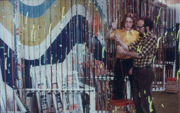 Jeune homme à chemise orange et homme à collier de barbe et lunettes dans un magasin de cannes à pêche. Traces de peinture jaune.