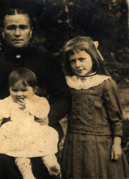 Femme avec fillette sur les genoux et petite fille debout à ses côtés en robe à col en dentelle et ruban dans les cheveux. Noir et blanc.