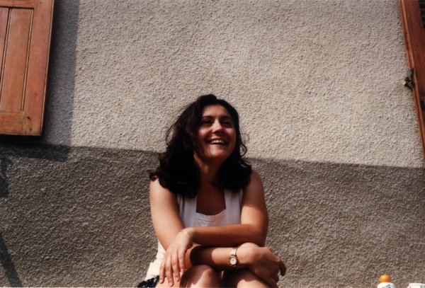 Femme brune souriante assise devant la façade d’une maison peinte en blanc et gris.