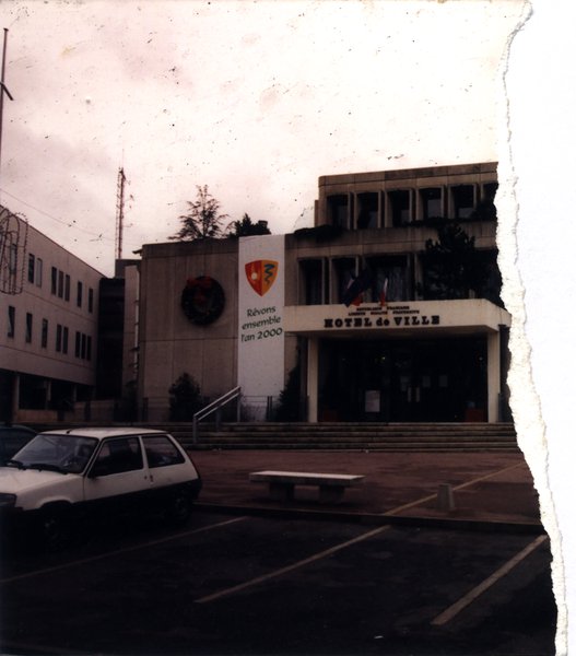 Voiture blanche garée sur une aire de stationnement devant une entrée. Fragment d’image déchirée.