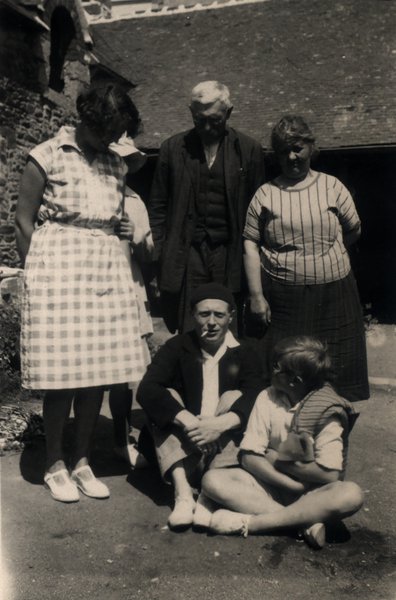 Famille de cinq personnes posant devant une maison en pierre. Noir et blanc.ᴬᴵ