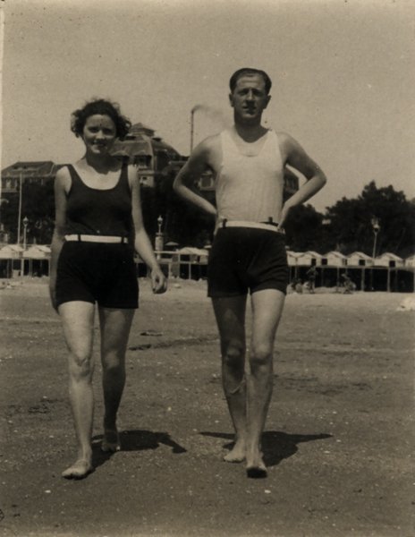 Homme et femme marchant sur une plage en tenue de bain, cabines en arrière-plan. Noir et blanc.ᴬᴵ