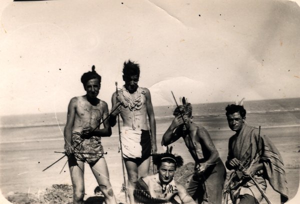 Groupe d'hommes déguisés avec plumes et lances sur une plage. Noir et blanc.ᴬᴵ