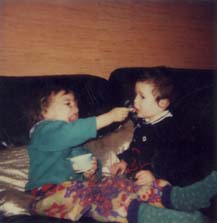 Petit enfant assis sur des coussins le visage barbouillé donne à manger à un autre enfant.