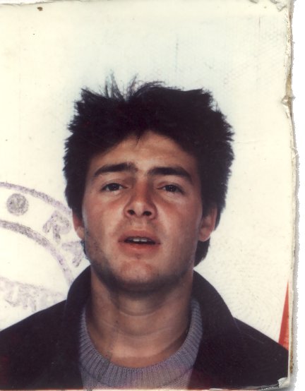 Visage d’un jeune homme brun avec veste et pull gris. Image avec trace de tampon encreur bleu.