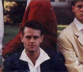 Jeune homme brun assis avec chemise blanche et veste bleu marine et deux autres jeunes hommes en veste blanche et rouge.