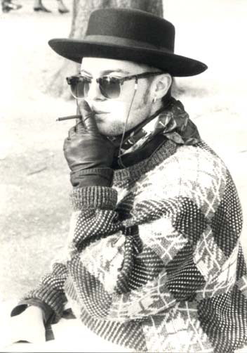 Homme en pull-over, chapeau, et lunettes noires, un petit cigare à la bouche. Noir et blanc.