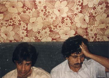 Deux hommes bruns assis sur un canapé, papier peint à grosses fleurs en arrière-plan.
