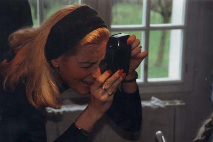 Femme blonde avec bandeau noir et ongles peints en rouge, l’œil sur l’objectif d’un appareil photo, fenêtre en arrière-plan.