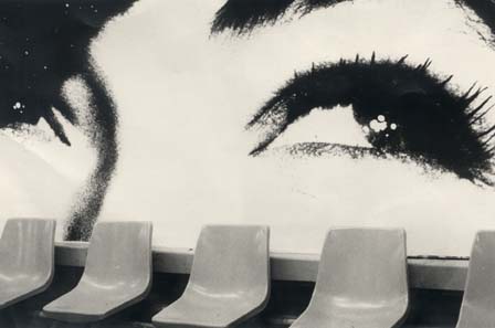Sièges en plastiques alignés, affiche avec grands yeux de femme en arrière-plan. Noir et blanc.