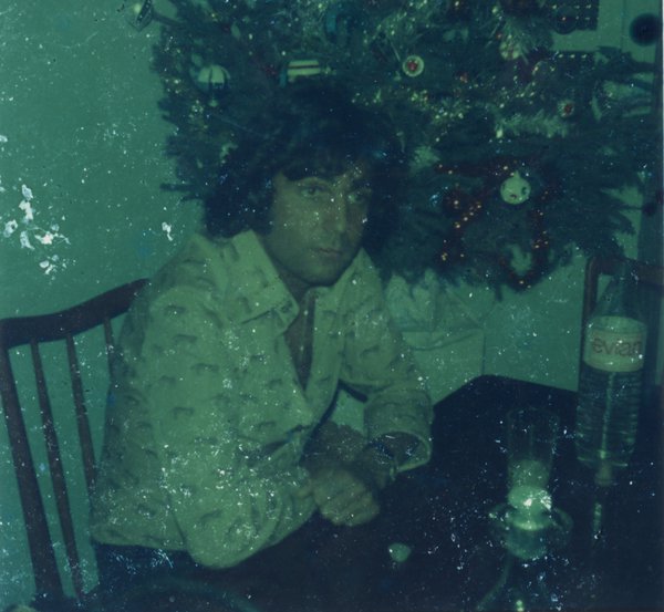 Homme brun chemise ouverte assis à une table devant un sapin de noël.