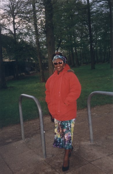 Femme souriante debout en anorak rouge sur un terre-plein devant une rangée d’arbres.
