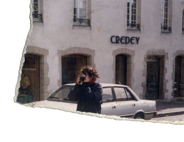 Femme l’œil sur le viseur d’un appareil photo devant une voiture blanche dans la rue. Fragment.