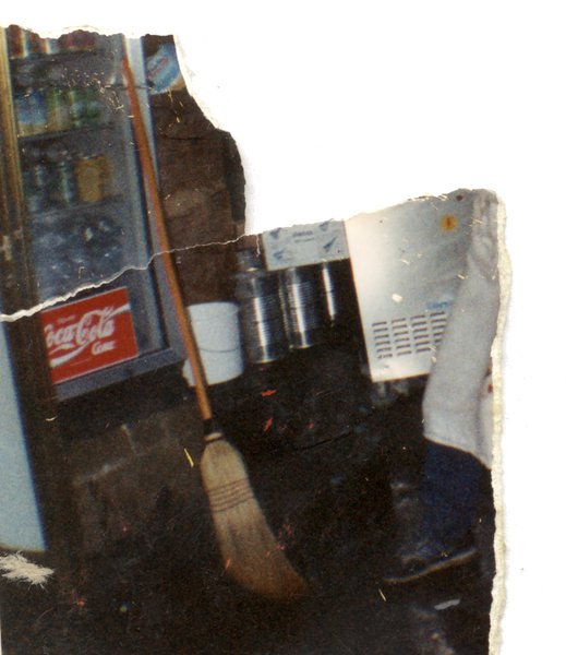 Balai posé devant un réfrigérateur à boissons conteant des packs de Coca-cola. Fragment.