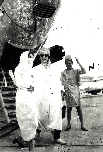 Trois personnes au pied d'un escalier d'avion, dont une hôtesse de l'air. Noir et blanc.ᴬᴵ