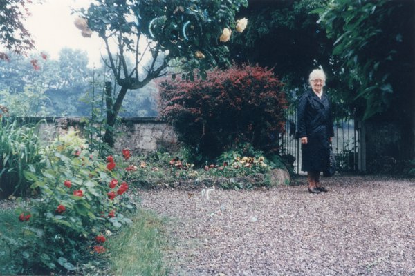 Femme à cheveux blancs debout sur le sol en gravier d’un jardin.