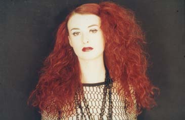 Femme  maquillée à grande tignasse rousse, le buste habillé d’un top en résille noire.