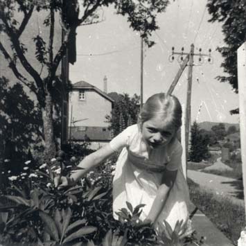 Petite fille blonde en robe blanche agenouillée dans la végétation, en arrière-plan poteaux électriques et une maison. Noir et blanc.