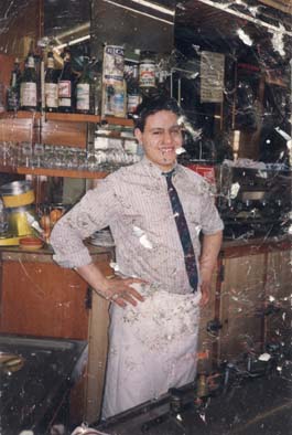 Homme souriant en bras de chemise et tablier blanc derrière un comptoir de café. Image abîmée.