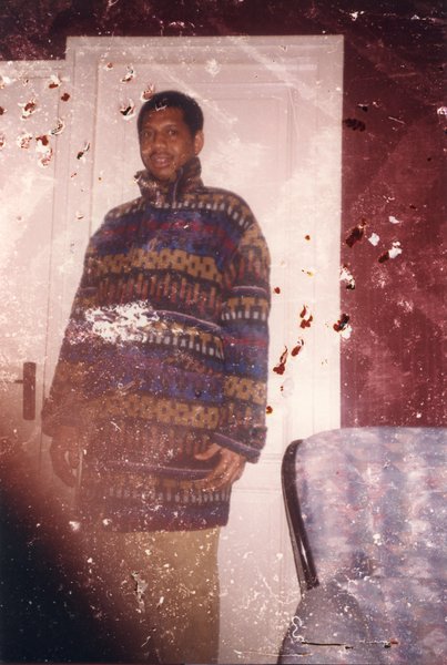 Homme en manteau bariolé debout devant une porte. Image avec traces de piqûres.