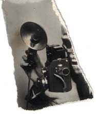 Appareil photo argentique et projecteur flash tenu à deux mains par un individu au visage caché par un masque de squelette. Fragment.