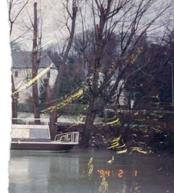 Bateau sur l’eau, bâtisses et rangée d’arbres en arrière-plan. Image avec impression date et heure. Fragment.