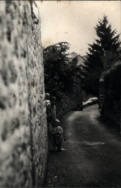 Mur où sont postés un homme, un chien et un petit enfant sur un chemin de terre fermé par un portail. Noir et blanc.