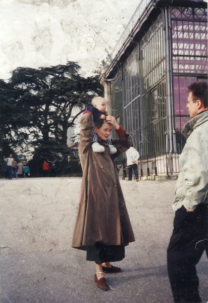 Femme en manteau avec enfant sur les épaules devant un des bâtiments en verre du jardin des Plantes, homme en avant-plan.