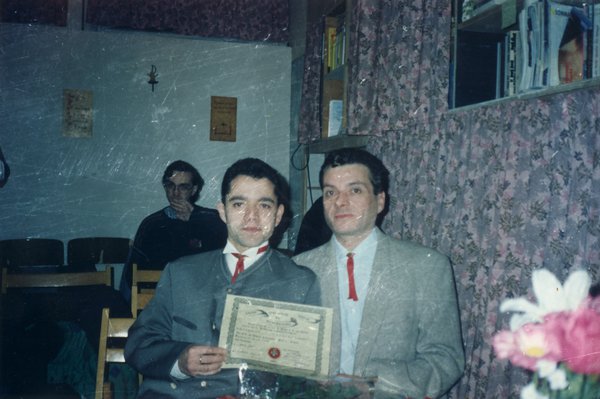 Deux hommes à lavallières rouges assis côte à côte à une table avec un diplôme dans les mains.