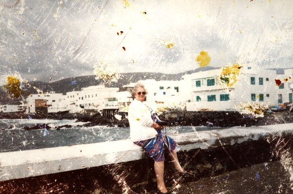 Femme âgée assise sur un muret face à la mer, village blanc en arrière-plan. Image très abîmée.ᴬᴵ