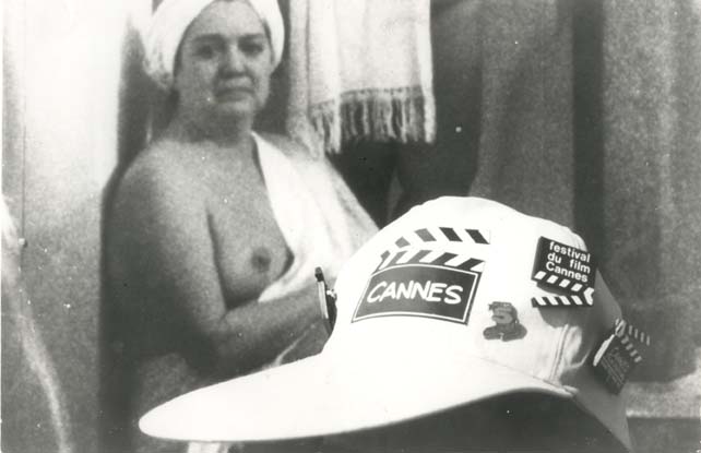 Casquette au logo du festival de Cannes, en arrière-plan une femme nue assise avec serviette éponge sur la tête et sur l’épaule. Noir et blanc.