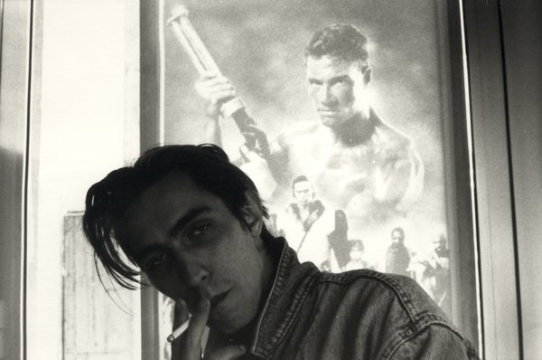 Homme à longue mèche tête penchée une cigarette à la bouche devant une affiche.
