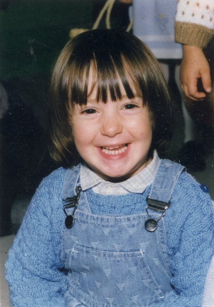 Petit enfant à frange brune et salopette en jean assis riant.