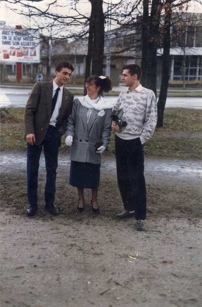 Femme à queue-de-cheval, lunettes, et gants blancs, entourés de deux jeunes hommes devant une rangée d’arbres.