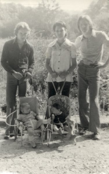 Deux hommes et une femme avec deux petits enfants dans deux poussettes, champ en arrière-plan.