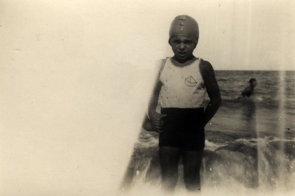 Garçon en bonnet et maillot de bain debout sur la plage, nageur dans la mer en arrière-plan. Noir et blanc.ᴬᴵ