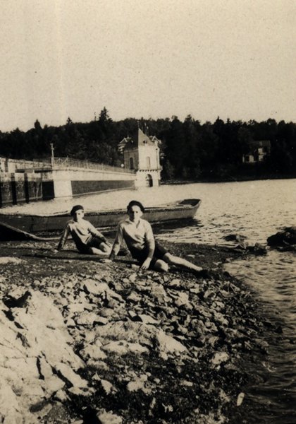 Deux femmes assises sur des rochers au bord d'un lac, bateau à l'arrière-plan. Noir et blanc.ᴬᴵ