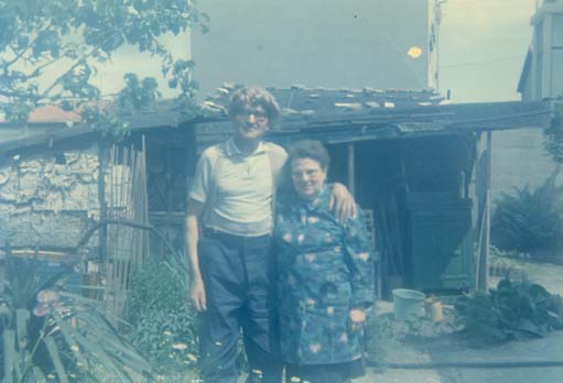 Homme âgé avec une perruque blonde sur la tête  tenant par l’épaule une femme âgée devant une cabane de jardin.