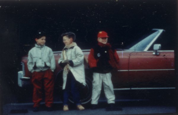 Trois enfants en manteaux debout devant une voiture rouge décapotable.