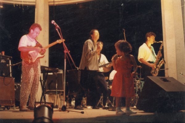 Groupe de musique sur scène en extérieur avec une enfant en robe rouge chantant.ᴬᴵ